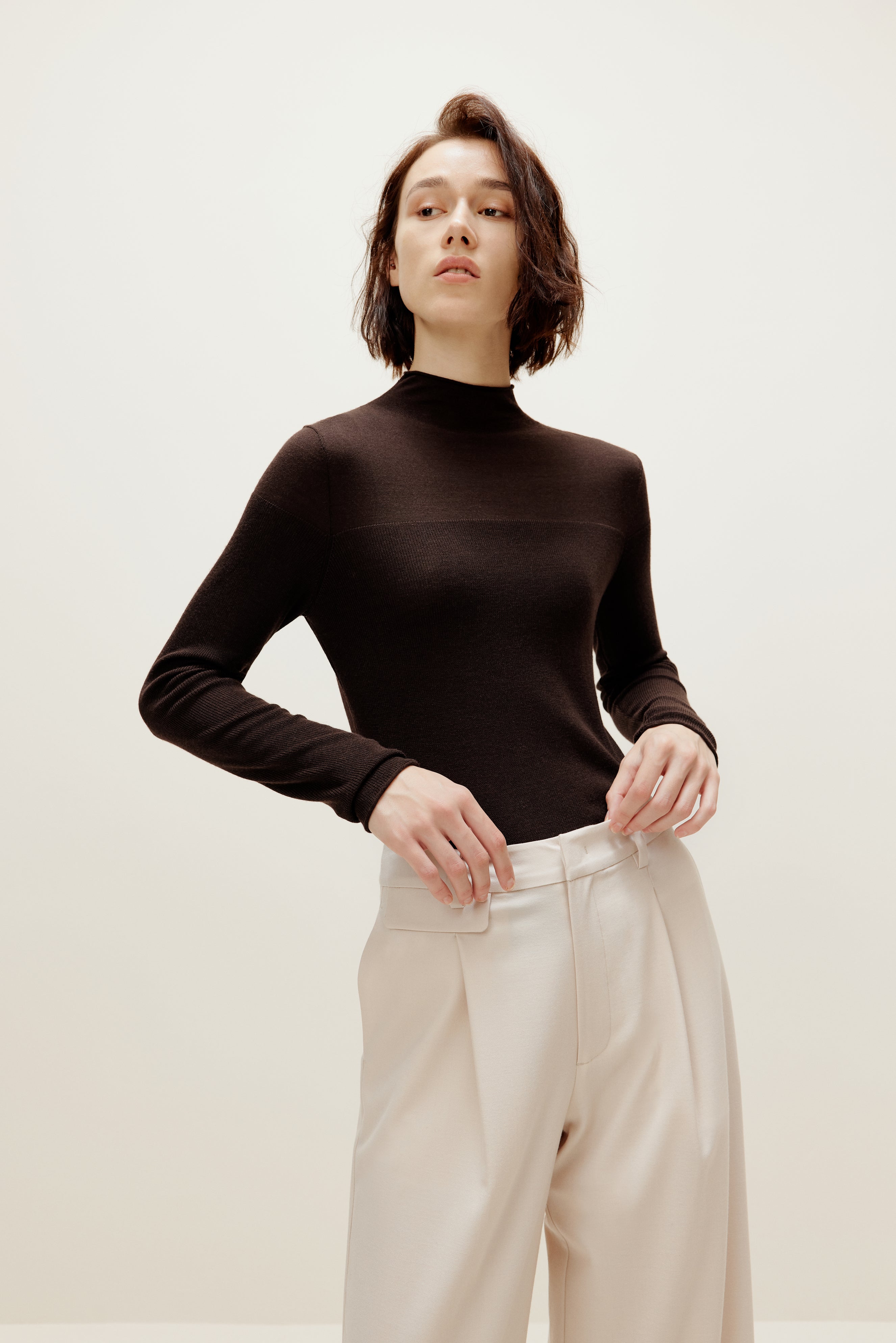 A woman wears a brown silky wool mock neck sweater and white pants.