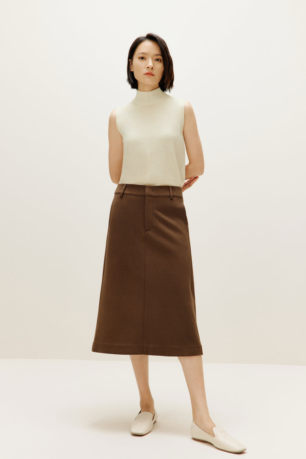 A woman wears a white silky wool mock neck sleeveless sweater and a brown skirt.