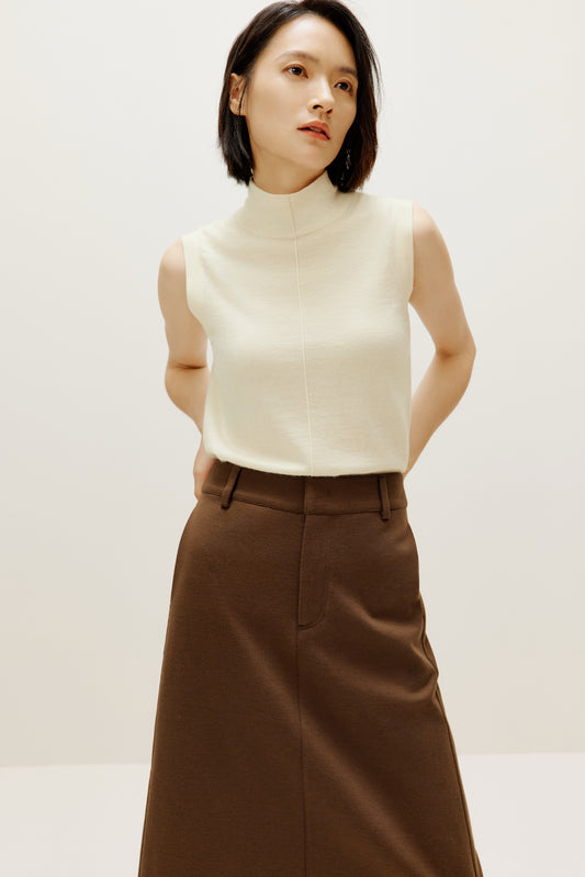 A woman wears a white silky wool mock neck sleeveless sweater and a brown skirt.