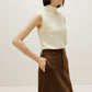 A woman wears a white silky wool mock neck sleeveless sweater and a brown skirt, facing right