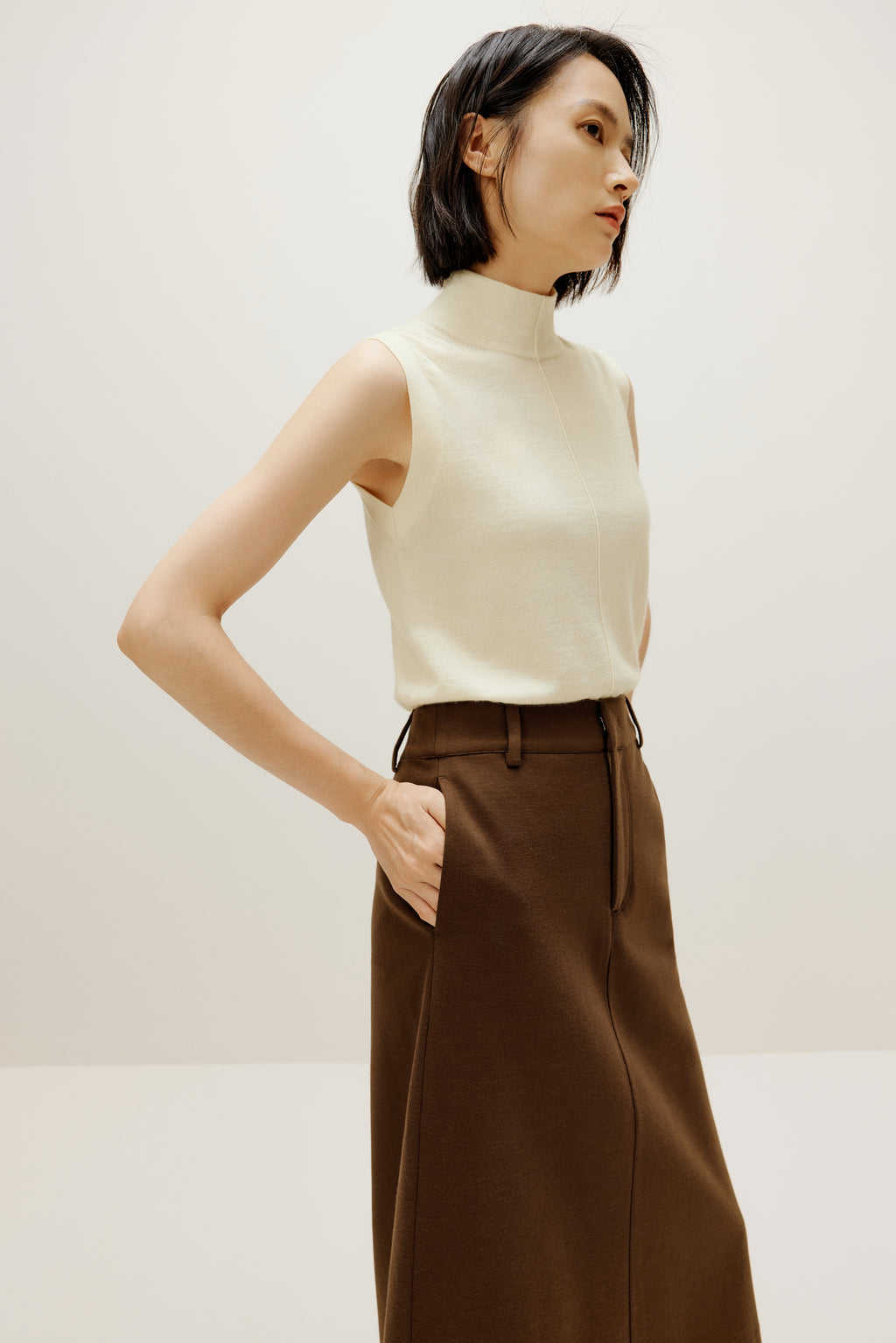 A woman wears a white silky wool mock neck sleeveless sweater and a brown skirt, facing right