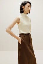 A woman wears a white silky wool mock neck sleeveless sweater and a brown skirt, facing right